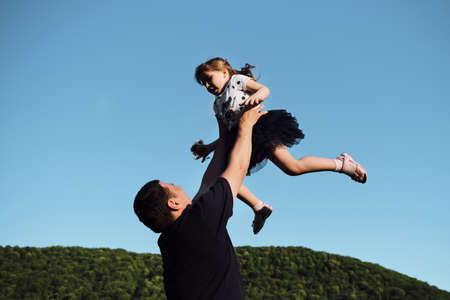 Spend time together on Fathers Day. Young Caucasian father turns his little daughter in his arms and laughs merrily together. Man throws little girl up against clear blue sky.の写真素材