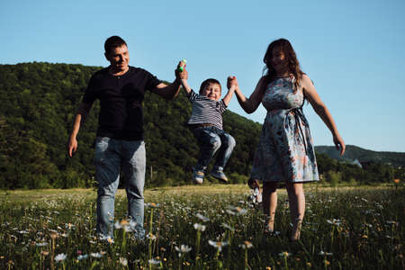 Young parents hold their young son with both hands and swing him back and forth and the child laughs merrily with his mouth wide open. Mom, dad and son on walk in chamomile field in summer.の写真素材