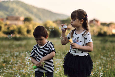 Children are standing side by side in chamomile field, blowing soap bubbles. Charming kids on walk in clearing relax among wild beautiful flowers. Happy childhood in nature.の写真素材