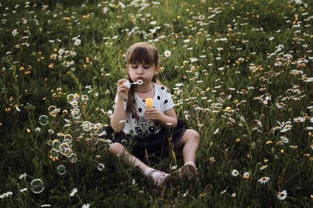 Little girl in dress sits in chamomile field and blows soap bubbles. Happy childhood in nature among flowers. Chamomile field and child at sunset.の写真素材