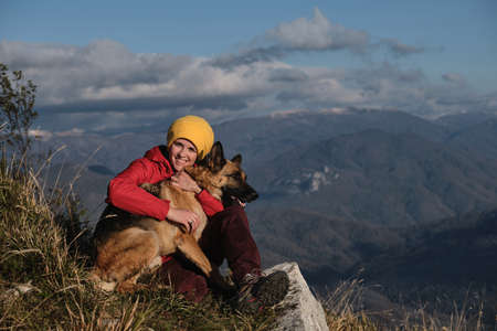 Young Caucasian woman in red jacket is sitting on top of mountain next to her dog and enjoying...