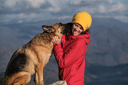Young pretty Caucasian woman in red jacket stands on top of mountain next to her dog and enjoys...