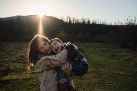 Caucasian boy in striped sweater is sitting merrily in arms of woman and they are both laughing hugging. Spend childhood in nature with beloved mother. Joy of motherhood. Warm setting sun.の写真素材