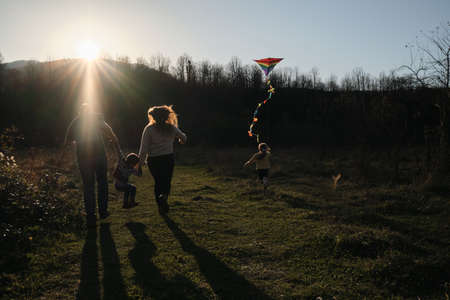 Young parents with son and daughter run across field at sunset and launch multicolored kite into sky. Have fun spending time with two children outdoors in nature. Rear view.の写真素材