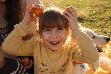 Portrait of funny little Caucasian girl in close up. Happy childhood. Child is sitting smiling and making imaginary ears on head with help of hands and two tangerines.の写真素材