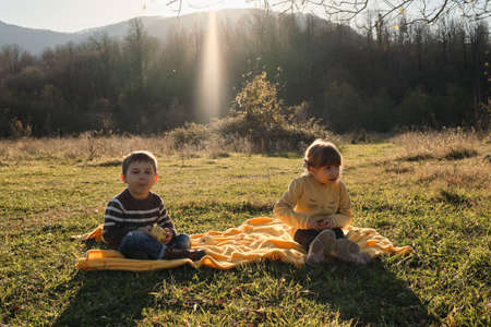 Little brother and sister are sitting in park at sunset on yellow blanket and eating fruit apples and tangerines. Charming Caucasian children. Spend childhood in nature.の写真素材