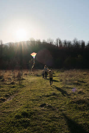 Have fun spending time with two children outdoors in nature. Joy of parenthood and motherhood. Mom walks on green grass with little son at sunset and daughter launches multicolored kite into sky.の写真素材