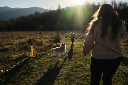 Mother with son and dog walk through field at sunset and launch multicolored kite into sky. Have fun spending time with child outdoors in nature. Rays of sun from behind mountain.の写真素材