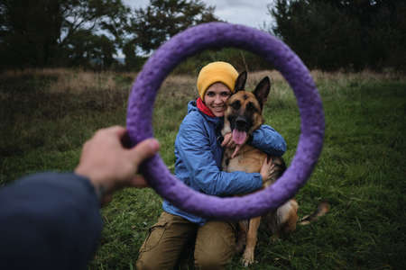 Young European white woman sits in green grass and hugs her German Shepherd dog. Have fun in park with furry human friend. Creative round dog toy frame.の写真素材