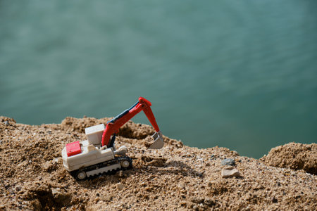 Construction and mining industry. creative banner. Small plastic toy excavator with bucket working on sand extraction at quarry. Pond in the background. Children's toy model of tractor extracts sand.の写真素材