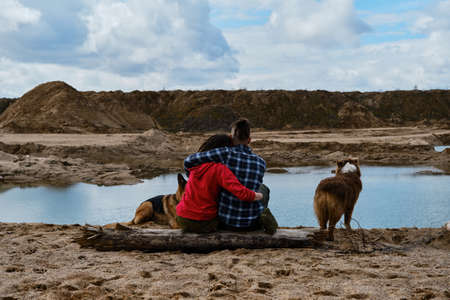 Young couple in love with dreadlocks sitting on wooden log on riverbank with two dogs and hugging...