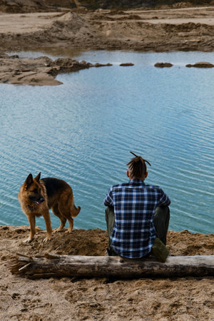 Young man with dreadlocks is sitting on wooden log on riverbank with dog enjoying views of nature. Sand pits and dunes and blue clear water. German shepherd with owner. rear view.の写真素材