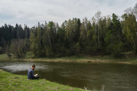 Young Caucasian man with dreadlocks and beard sitting in spring park on green grass riverbank and intently reading book completely immersed in another reality. Concept learning outside and lifestyle.の写真素材