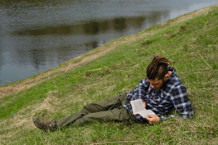 Young Caucasian man with dreadlocks and beard lying in spring park on green grass riverbank and intently reading book completely immersed in another reality. Concept learning outside and lifestyle.の写真素材