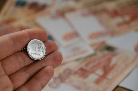 Human holds iron coin 1 ruble with hand on blurry background of five thousand paper bills. Economics...
