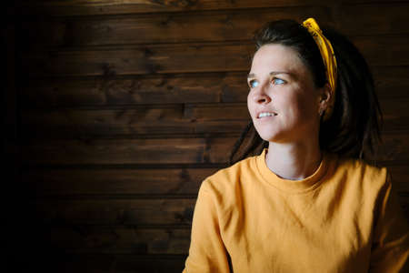 The concept of happiness and fun. Portrait of young Caucasian woman with dreadlocks in yellow hoodie and bandana on head against wooden wall. She smiles and looks away with blue eyes.の写真素材