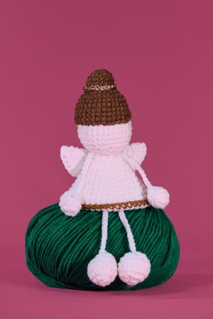 Amigurumi one white angel without face wears brown hat, sits on green ball of thread on pink background. A soft DIY toy made of natural cotton and wool. Cute little angel crocheted, handmade art.の写真素材