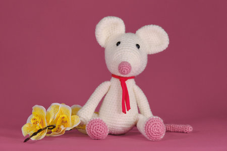 Amigurumi one white mouse with big ears and long nose sits on pink background next to yellow orchid flowers. A soft DIY toy made of natural cotton and wool. Cute little rat crocheted, handmade art.の写真素材