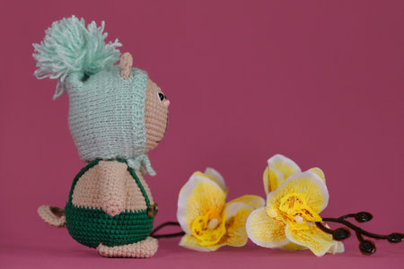 One brown cat wearing green jumpsuit and funny fluffy crocheted hat, handmade art. Amigurumi kitten doll on pink background next to yellow orchid flowers. A soft DIY toy made of cotton.の写真素材