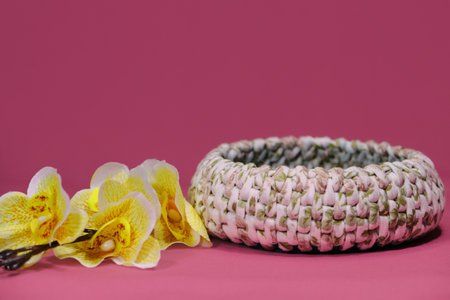 Crochet Home Basket on pink background. Green and white round box for small things made of thread crocheted, handmade art. Nearby is a branch of yellow orchid flowers. front view.の写真素材