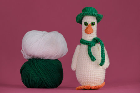 One white goose with orange beak wears green hat and scarf, crocheted from natural wool and cotton, handmade art. Amigurumi duck doll on pink background. Soft DIY toy made of cotton. front view.の写真素材