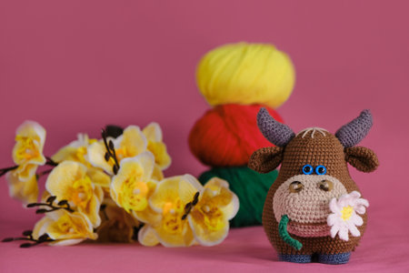 Amigurumi cow doll on pink background next to yellow orchid flowers and multicolored wool balls. A soft DIY toy made of cotton. One brown bull with daisy in mouth crocheted, handmade art.の写真素材
