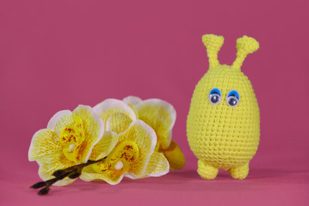 Freak humanoid crocheted, handmade art. Amigurumi one small yellow alien with horns and blue eyes stands on pink background next to yellow orchid flowers. Soft DIY toy made of natural cotton and wool.の写真素材