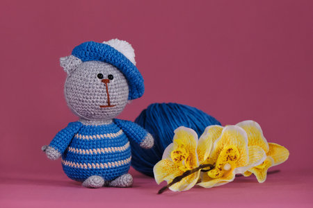 Amigurumi kitten doll on pink background next to yellow orchid flowers. A soft DIY toy made of cotton. One grey cat wearing blue striped clothes and artists hat crocheted, handmade art.の写真素材
