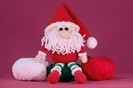 Amigurumi one Christmas gnome with red hat and white beard sits on pink background. Soft DIY toy made of natural cotton and wool. Crocheted santa claus, handmade art. Santas helper.の写真素材