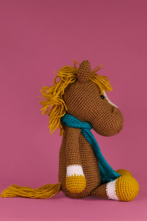 Cute little horse with scarf around neck crocheted, handmade art. Amigurumi one brown pony with orange mane sitting on pink background. Soft DIY toy made of natural cotton and wool. Side view.の写真素材