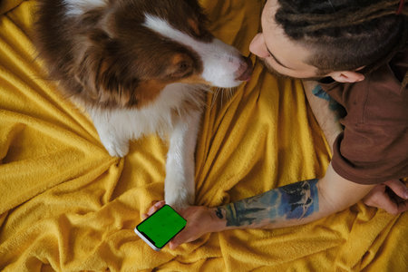 Young man lying on yellow blanket with Australian Shepherd dog, holding phone in hands and surfing Internet online top view. Green chroma Mockup Screen. Copy space for advertising pet shops.の写真素材