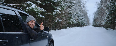 Young adult attractive Caucasian man sits at the wheel of his car snowy winter day showing thumbs up gesture. Wintertime road trip. Happy smiling hipster guy spitting in car and looking windowの写真素材