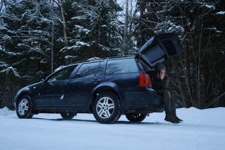 Young Caucasian man sits in the trunk of a car and using phone calling a mechanic for help. A broken-down vehicle in a snowy winter forest. An empty rural road. Rescue service is requiredの写真素材