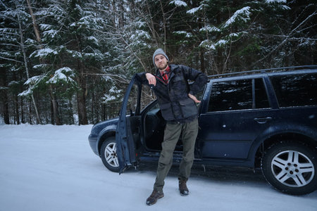 A young Caucasian man stands with keys in his hands near the car. The guy is smiling, he just bought a new car. Snowy winter forest and the owner of the vehicle. Leasing of a used car. front viewの写真素材