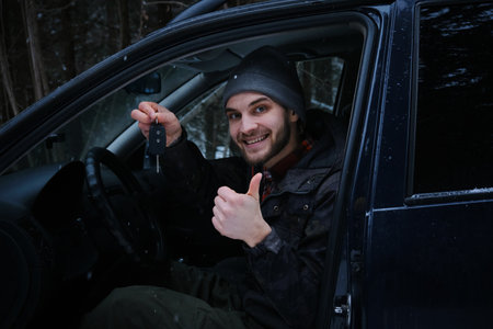 Caucasian man is sitting in a car with a black interior and keys in his hands. The guy is smiling, he just bought a new car. Snowy winter forest and the owner of the vehicle. Leasing of a used autoの写真素材