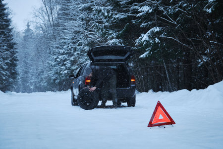 The sign is a red triangle on a rural winter snowy forest road. A broken-down car after the incident. The man took out a spare tire to change itの写真素材