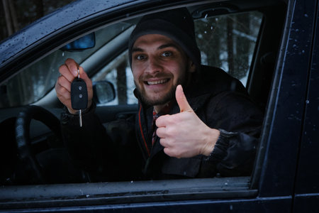 Caucasian man is sitting in a car with a black interior and keys in his hands. The guy is smiling, he just bought a new car. Snowy winter forest and the owner of the vehicle. Leasing of a used autoの写真素材