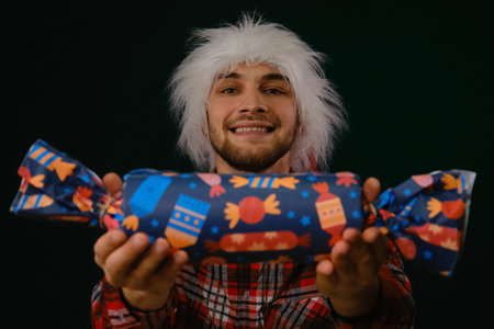 Concept of Happy New Year and Merry Christmas. Minimalistic festive studio dark background with one person. Young Caucasian guy wears a red Santa hat and holds a big candy surprise gift in handsの写真素材