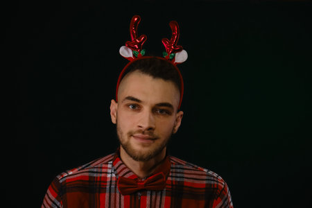 Concept of New Year and Christmas. Minimalistic festive dark studio background with one person. Caucasian guy wears a reindeer horn headband, a plaid shirt and a bow tie. Emotion of joy and happinessの写真素材