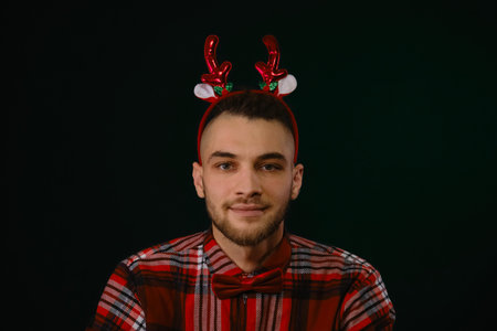 Concept of New Year and Christmas. Minimalistic festive dark studio background with one person. Caucasian guy wears a reindeer horn headband, a plaid shirt and a bow tie. Emotion of joy and happinessの写真素材