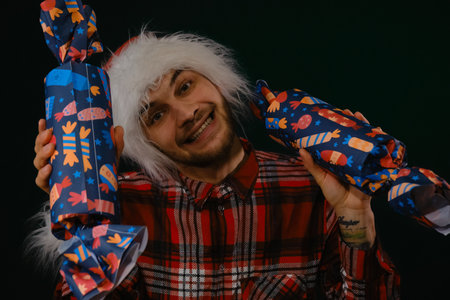 Concept of New Year and Christmas. Minimalistic festive studio background with person. Young Caucasian guy wears a red Santa hat and holds large candy gifts in hands. Emotions of happiness and joyの写真素材