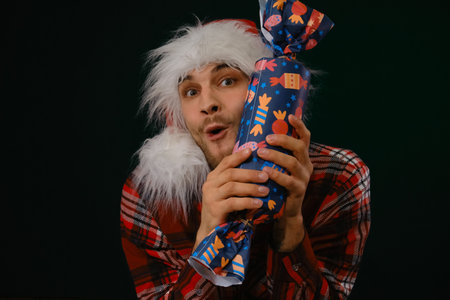 Concept of Happy New Year and Merry Christmas. Minimalistic festive studio dark background with one person. Young Caucasian guy wears a red Santa hat and holds a big candy surprise gift in handsの写真素材