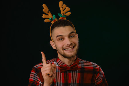 Concept of Happy New Year and Merry Christmas. A young Caucasian guy wears a reindeer horn headband, a plaid shirt and a bow tie. Gesture idea - index finger up. Front view close up portraitの写真素材