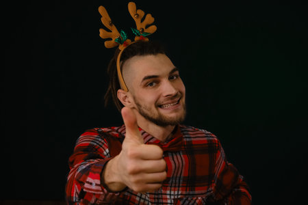 Happy New Year and Merry Christmas. Caucasian guy wears a reindeer horn headband, a plaid shirt and a bow tie. Gesture of approval and like a thumbs up. Front view portrait. Dark studio backgroundの写真素材