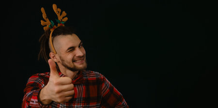 Happy New Year and Merry Christmas. Caucasian guy wears a reindeer horn headband, a plaid shirt and a bow tie. Gesture of approval and like a thumbs up. Dark studio banner with copy spaceの写真素材