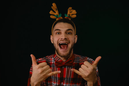 Happy New Year and Merry Christmas. A young Caucasian guy wears a reindeer horn headband, a plaid shirt and a bow tie. Santa's reindeer helper. Gesture greeting surfers. Emotions of happiness and joyの写真素材
