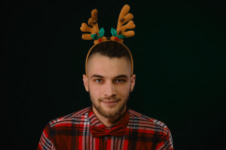 New Year and Christmas. Minimalistic studio background with person. Caucasian guy wears a reindeer horn headband, a plaid shirt and a bow tie. A piercing, mesmerizing look from under his browsの写真素材
