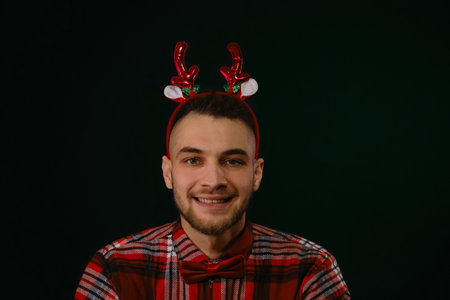Concept of New Year and Christmas. Minimalistic festive dark studio background with one person. Caucasian guy wears a reindeer horn headband, a plaid shirt and a bow tie. Emotion of joy and happinessの写真素材
