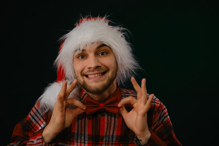Concept of New Year and Christmas. Minimalistic festive studio background with person. Caucasian guy wears a red hat, a plaid shirt and a bow tie. Emotions of happiness and joy. Front view portraitの写真素材
