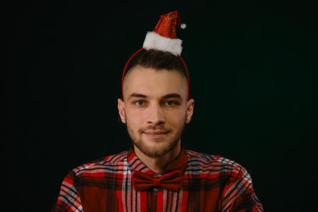 Concept of New Year and Christmas. Minimalistic festive studio background with person. A young Caucasian guy wears a red Santa hat headband, a plaid shirt and a bow tie. Emotions of happiness and joyの写真素材